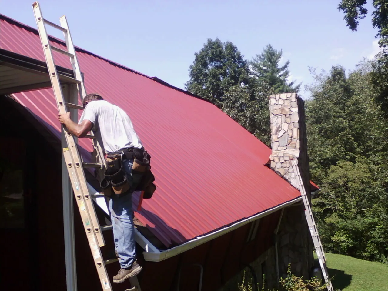 Roofer on a ladder beside a red metal roof and stone chimney — Soffit & Fascia Repair by Roofing Brothers in Lopez Island